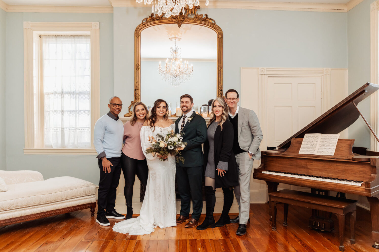 Elopement Wedding A bride in a white dress and groom in a dark suit stand in the center of a room with four other people, two on each side, all smiling. They are in a well-lit room with a grand piano on the right and a large mirror behind them. The floor is wooden, and there's a white couch on the left. Elopements Inc