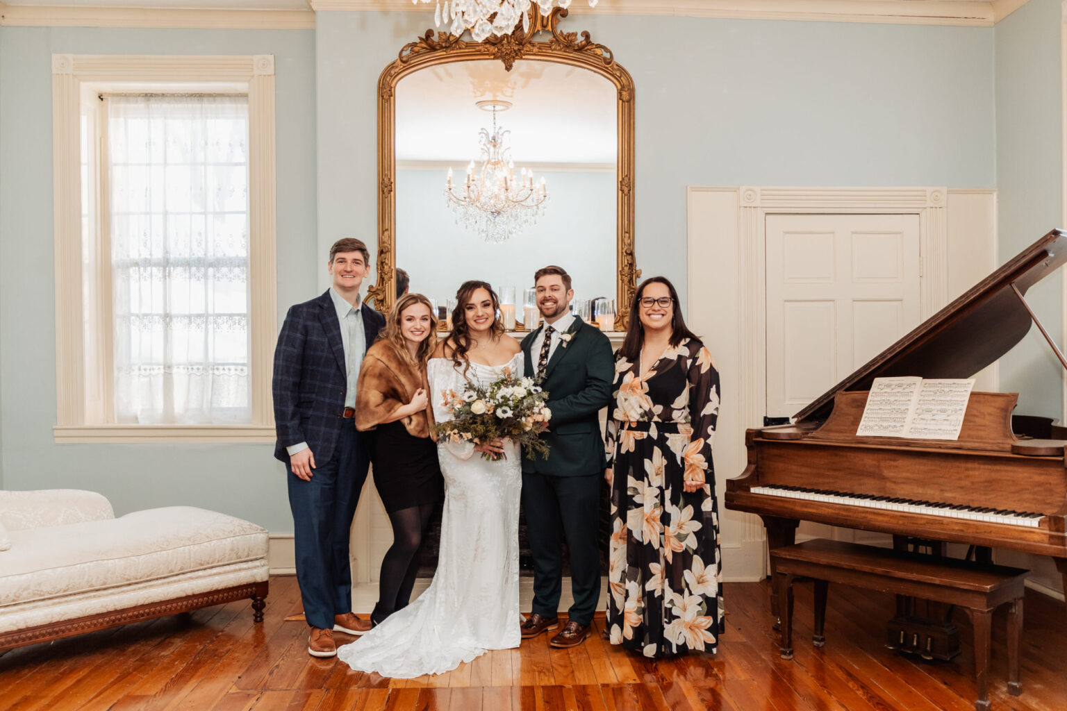 Elopement Wedding A group photo featuring five people standing in an elegant room. The group includes a bride in a white dress, holding a bouquet, and a groom in a dark green suit. They are flanked by two women, one in a black floral dress and the other in a fur stole, and a man in a blue suit. There's a grand piano to the right with sheet music, a chandelier above, a large mirror, light blue walls, and a cushioned bench to the left. Elopements Inc