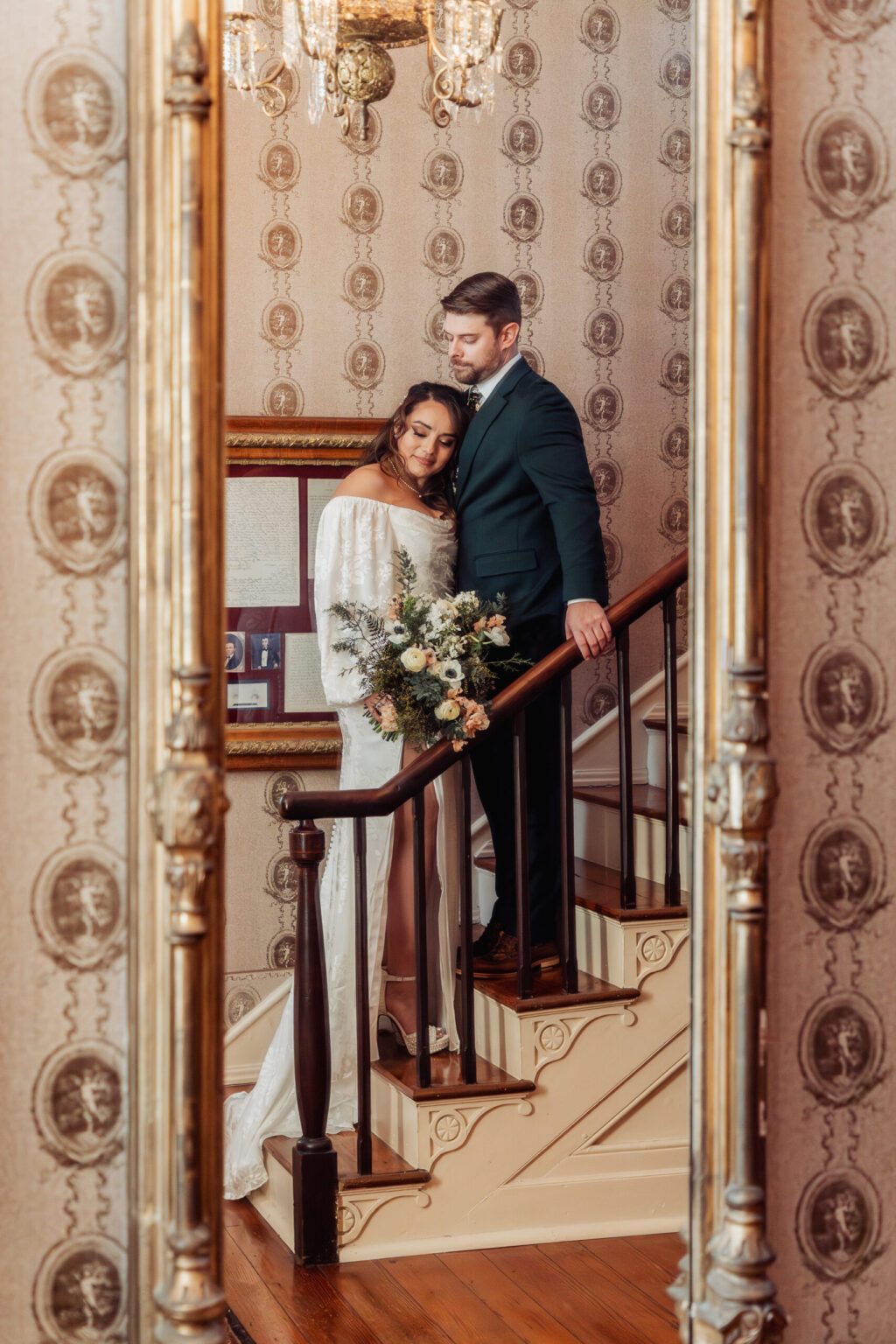 Elopement Wedding A couple stands on a wooden staircase with vintage wallpaper, the woman in a white off-shoulder dress holding a bouquet, and the man in a dark suit leaning against the bannister. They are reflected in a large ornate mirror, with a chandelier hanging above them. The setting is elegant and historic. Elopements Inc