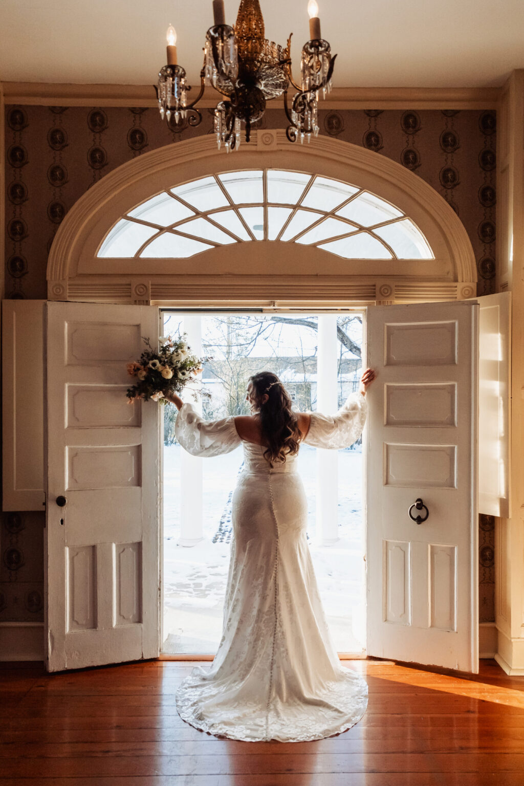 Elopement Wedding A bride in a long, white lace gown stands in an arched doorway, framed by ornate double doors with glass panels. She holds a bouquet of flowers and looks outside to a snowy scene. The room features elegant wallpaper, a chandelier, and wooden floors, illuminated by soft natural light. Elopements Inc