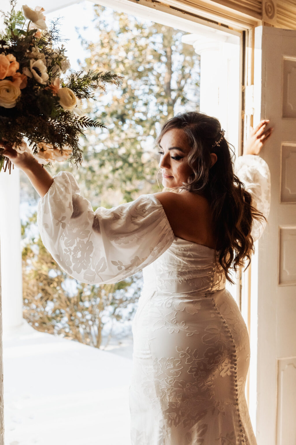 Elopement Wedding A bride wearing a lace off-the-shoulder wedding dress stands at an open door, bathed in natural light. She has long, wavy hair and holds a bouquet of flowers with greenery in her right hand. She looks downward with a serene expression, and the background shows a snowy outdoor scene. Elopements Inc