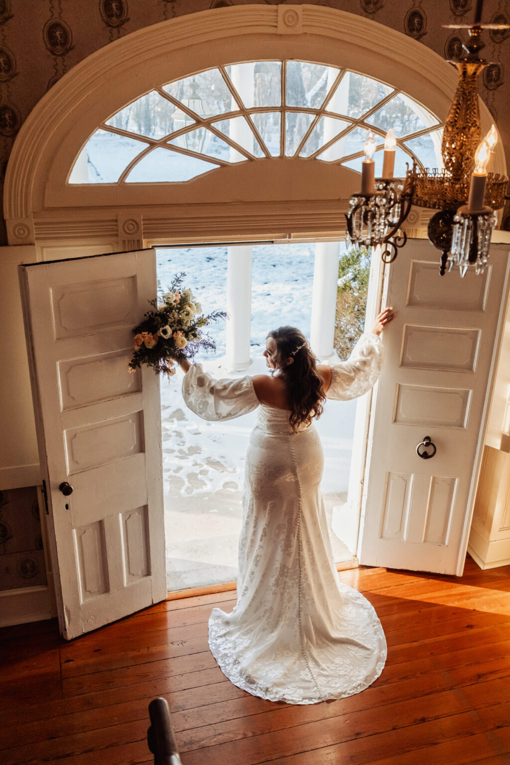 Elopement Wedding A bride in a white, long-sleeved wedding gown stands in front of open double doors with curved transom windows, looking outside. She holds a bouquet of flowers in her left hand. Sunlight filters through, casting soft shadows. A chandelier hangs from the ceiling above the wooden floor. Elopements Inc