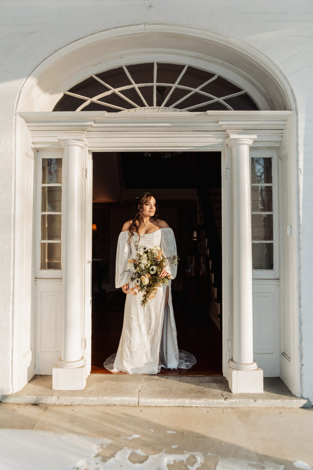 Elopement Wedding A bride in a flowing white gown with long, translucent puff sleeves stands in an arched doorway of a white building. She holds a bouquet of white and green flowers. Sunlight casts soft shadows, illuminating her and the classic architectural details surrounding the doorway. Elopements Inc