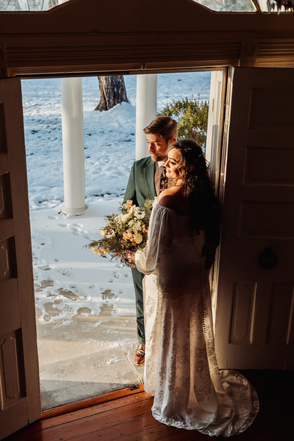 Elopement Wedding A couple stands in an open doorway, warmly lit. The bride, in an off-shoulder white gown with lace details, holds a bouquet of flowers. The groom, in a green suit, stands beside her. Outside, snow covers the ground, and the scene exudes a serene, wintery atmosphere. Elopements Inc