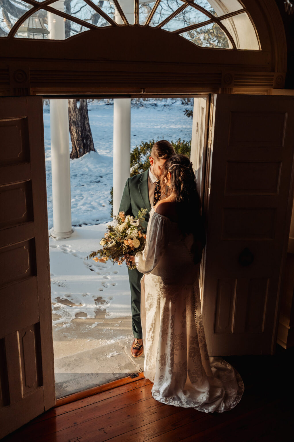 Elopement Wedding A bride and groom share a kiss while standing in a doorway. The bride, in a lace gown, holds a bouquet of white and pastel flowers. The groom wears a dark green suit. Behind them, sunlight illuminates the snowy landscape outside, with trees and columns visible. Elopements Inc