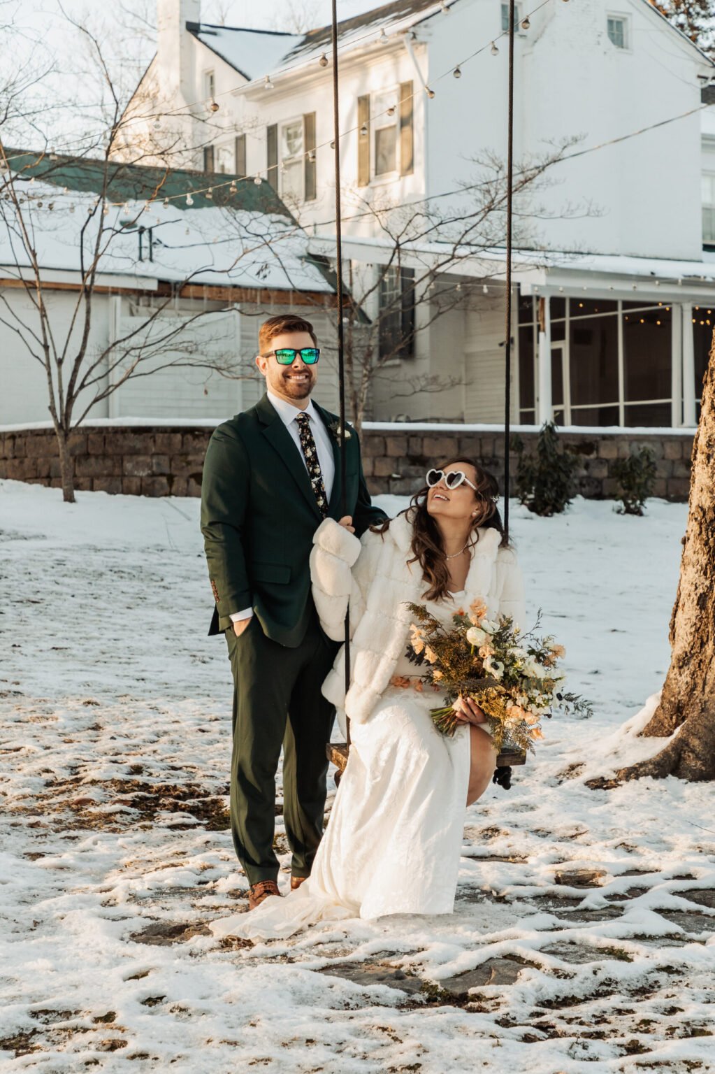 Elopement Wedding A couple stands outdoors in a snowy setting. The woman is seated on a swing, wearing a white wedding dress, a fur shawl, and sunglasses, while holding a bouquet of flowers. The man, dressed in a green suit and sunglasses, stands beside her with one hand on the swing rope. A house is in the background. Elopements Inc