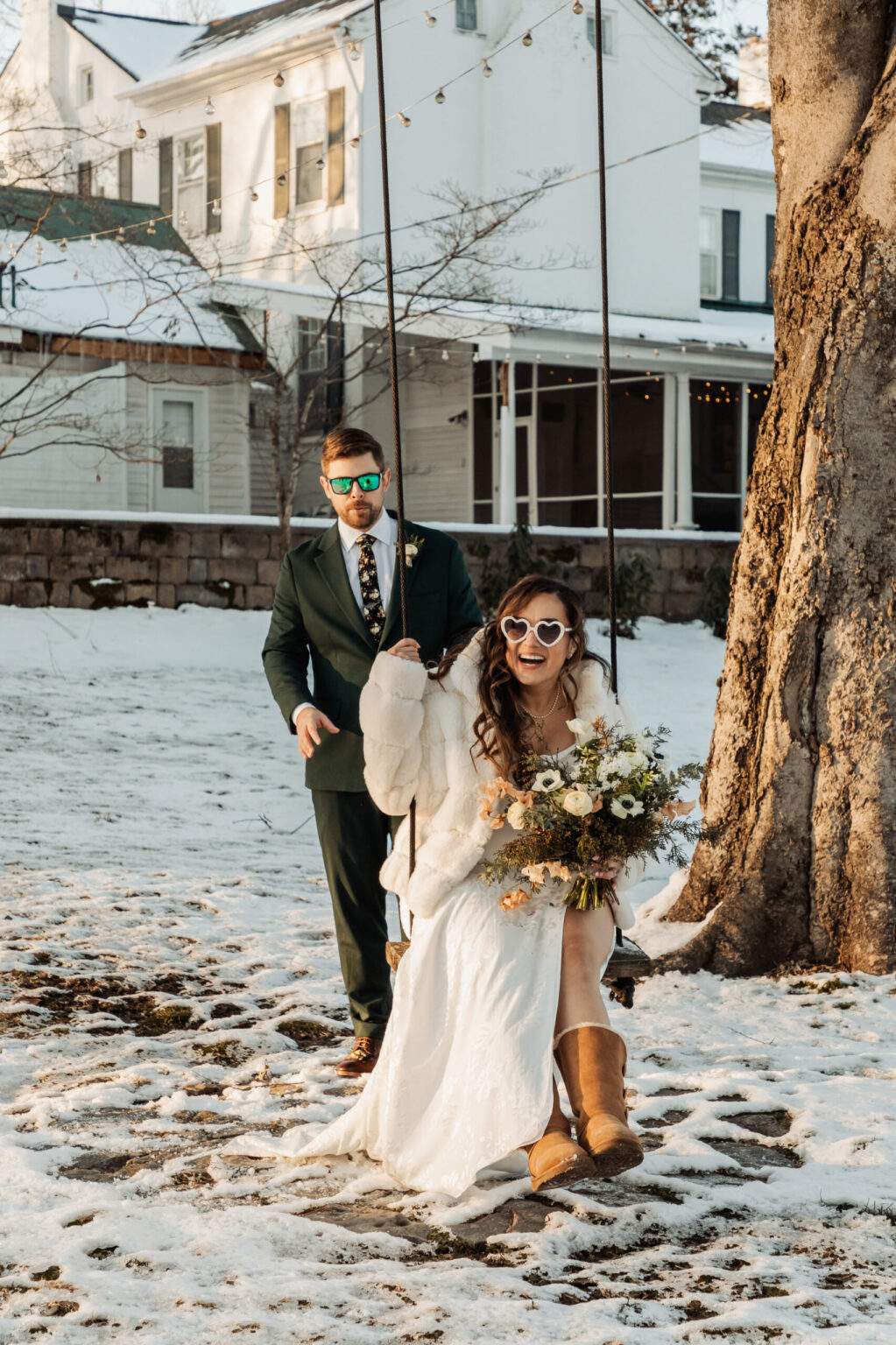 Elopement Wedding A bride in a white dress, sunglasses, and brown boots sits on a swing holding a bouquet, smiling. A groom in a green suit and sunglasses stands behind her, holding her white fur wrap. They are outside a snow-covered house with a tree adorned with string lights. Elopements Inc