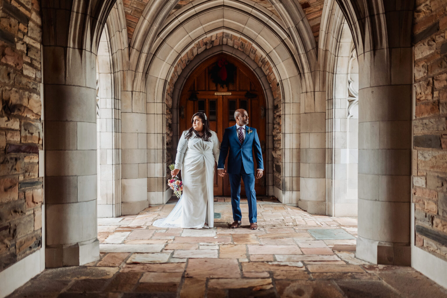 Elopement Wedding A bride and groom stand under an ornate stone archway. The bride, in a white gown and holding a bouquet, gazes to her left. The groom, in a blue suit and tie, looks to his right. They hold hands, both with serious expressions. The floor is made of irregular stone tiles. Elopements Inc