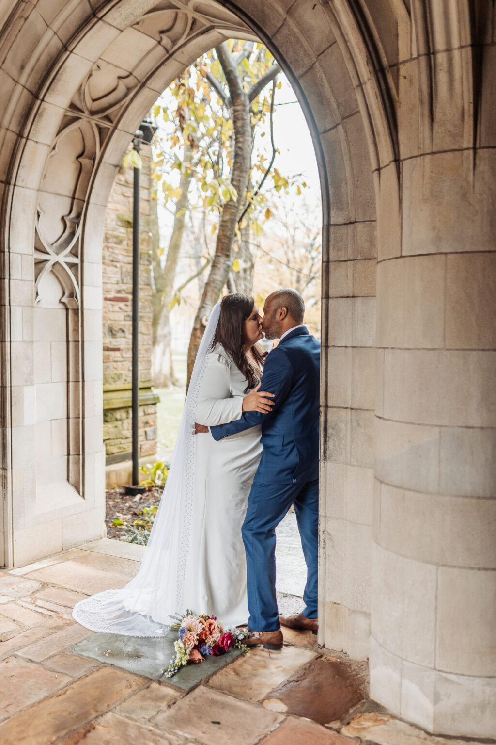 Elopement Wedding A couple kisses under an arched stone doorway. The bride, in a white gown with a long veil, stands next to the groom, who is wearing a blue suit. A bouquet of flowers is placed on the ground nearby. The scene appears to be in an outdoor stone courtyard with autumn trees in the background. Elopements Inc