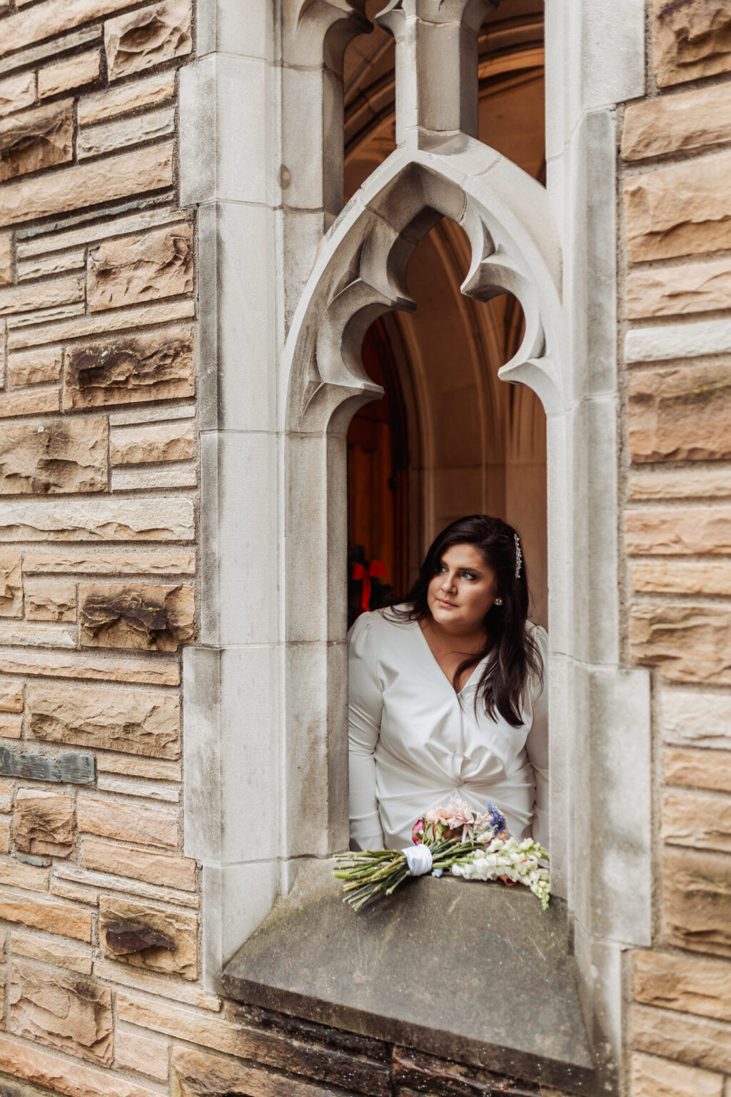 Elopement Wedding A woman in a white dress stands at a narrow, arched window made of stone bricks. She gazes to the side, holding a bouquet of flowers, with her arms resting on the windowsill. The window's gothic-style design adds an architectural elegance to the scene. Elopements Inc