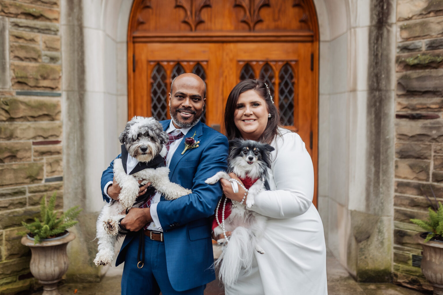 Elopement Wedding A happy couple stands outside a stone building with wooden doors, each holding a dog. The man on the left, wearing a blue suit and tie, holds a small black and white dog. The woman on the right, in a white dress, holds a small black and white fluffy dog dressed in a red outfit. Elopements Inc