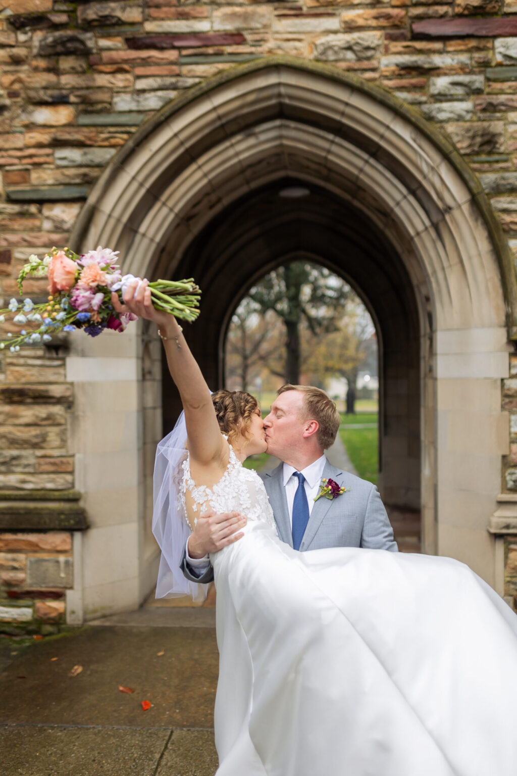 Elopement Wedding A bride and groom share a kiss in front of a stone archway. The groom, wearing a light gray suit, holds the bride in his arms as she raises a colorful bouquet. The bride is dressed in a white wedding gown with a lace bodice and veil. The background features a stone building and greenery. Elopements Inc