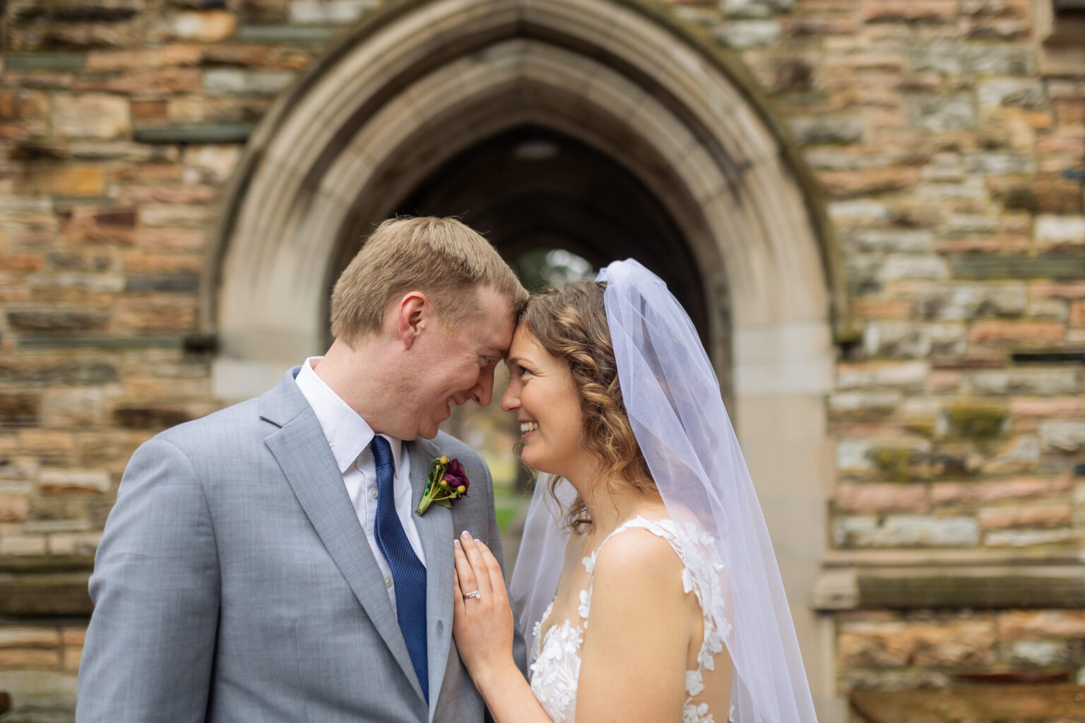 Elopement Wedding A bride and groom share a joyful moment on their wedding day, touching foreheads and smiling. The groom wears a gray suit with a blue tie and boutonniere. The bride wears a white wedding dress with a lace top and a veil. They are standing in front of a stone archway. Elopements Inc