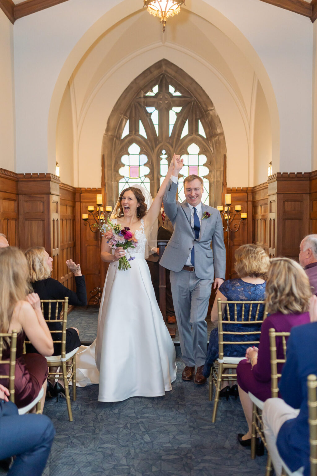 Elopement Wedding A bride and groom joyfully hold hands and raise their arms in celebration as they walk down the aisle of a church. The bride wears a white gown and holds a bouquet of flowers, while the groom is in a blue suit and tie. Guests seated on both sides applaud. The venue features wooden pews and arched windows. Elopements Inc