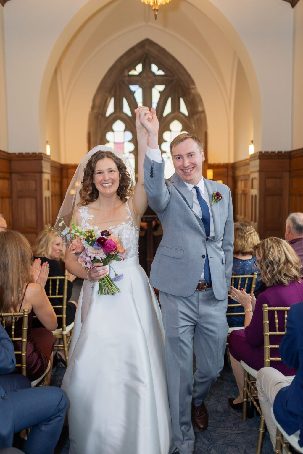 Elopement Wedding A bride in a white dress and veil and a groom in a light gray suit joyfully raise their clasped hands while walking down the aisle. The bride holds a bouquet of pink and purple flowers. They are smiling broadly, and guests are seated on either side, clapping and celebrating the couple. Elopements Inc