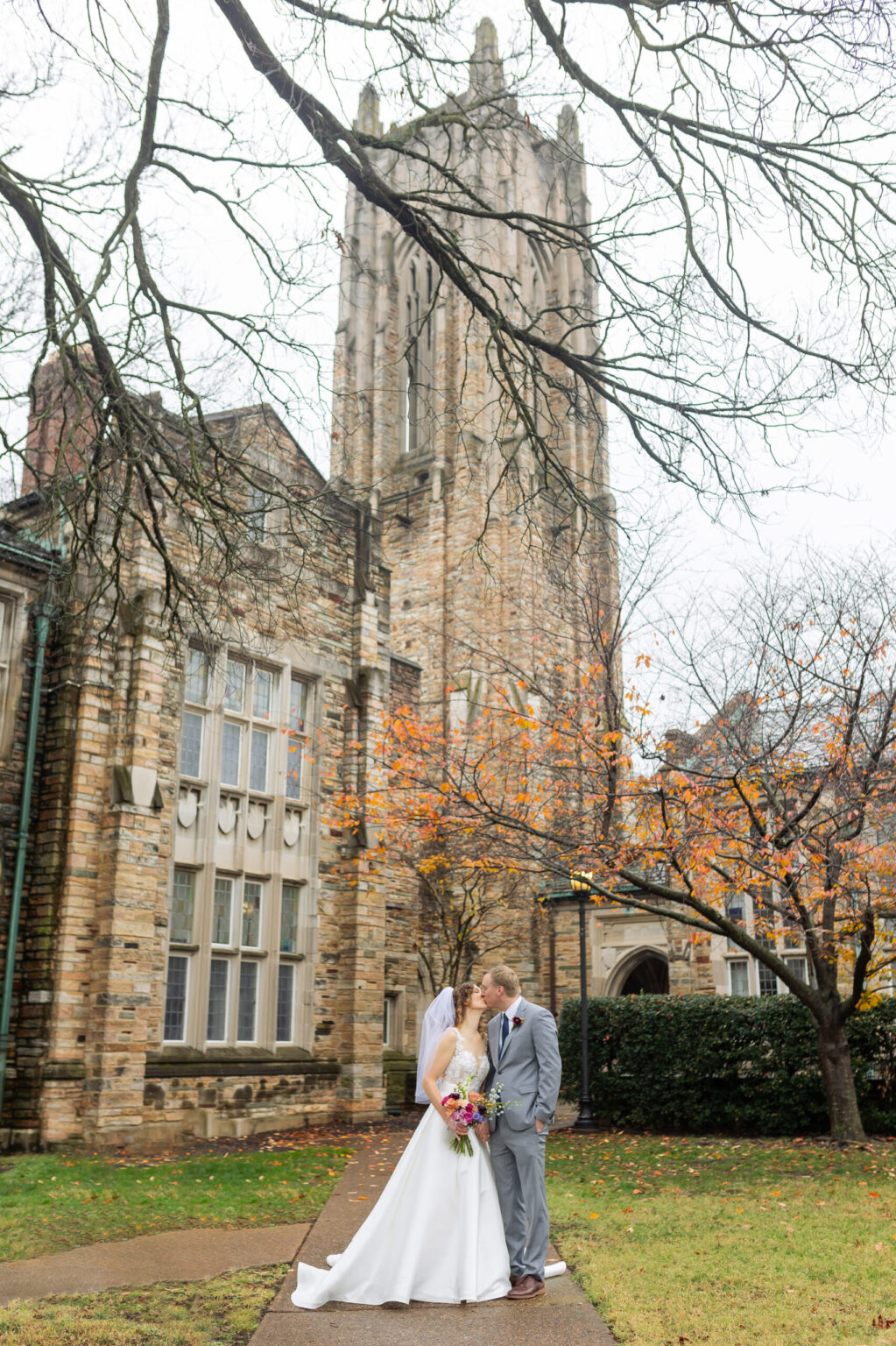 Elopement Wedding A bride and groom share a kiss on a path beside a historic stone building with tall, detailed architecture. The bride wears a white dress and veil, holding a bouquet, while the groom is in a light gray suit. Bare trees and autumn leaves add a seasonal touch to the scene. Elopements Inc