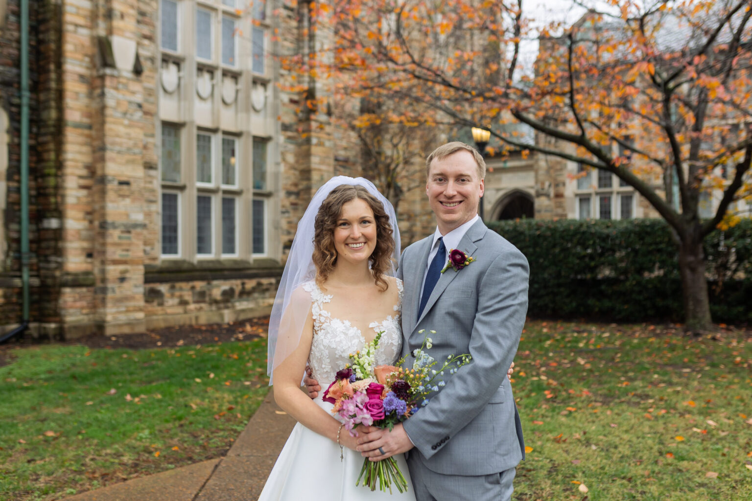 Elopement Wedding A smiling couple stands outdoors in front of a stone building with large windows and fall foliage. The bride wears a white dress with a veil, holding a bouquet of vibrant flowers. The groom is in a light gray suit with a purple boutonniere. The ground is wet, suggesting recent rain. Elopements Inc