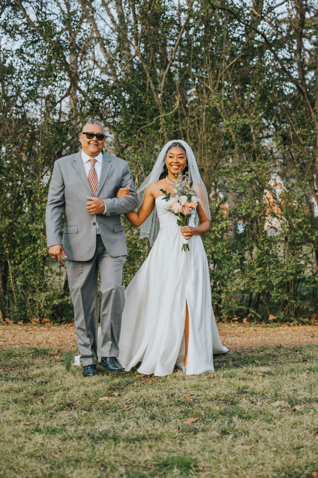 Elopement Wedding A bride in a white strapless wedding dress and veil walks into an outdoor ceremony while holding a bouquet of flowers. She is accompanied by a man in a gray suit and sunglasses. They are walking on grass with a backdrop of trees and foliage. Both appear happy and smiling. Elopements Inc