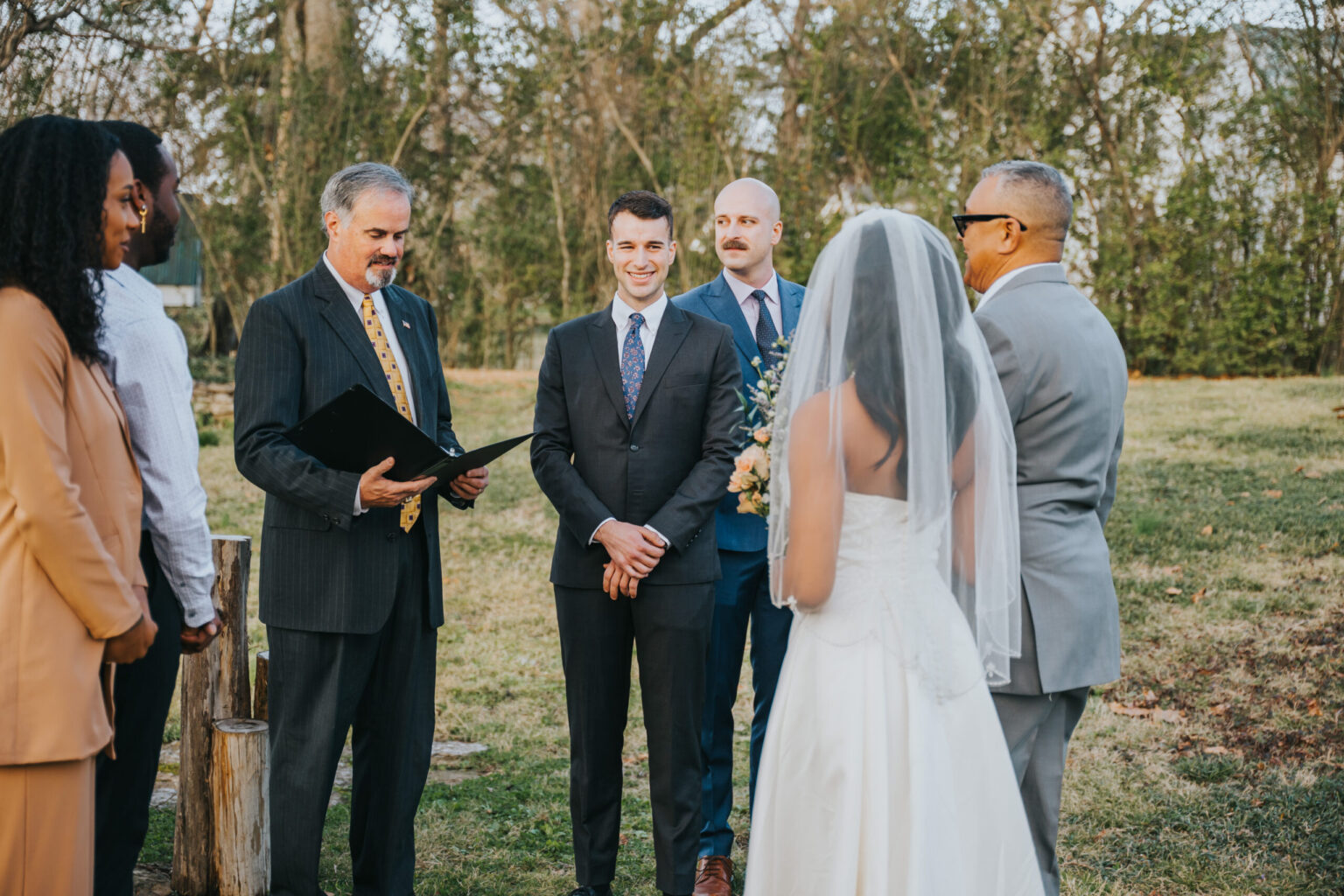 Elopement Wedding A wedding ceremony outdoors with six people visible. A bride in a white dress and veil stands facing a groom in a dark suit. A person officiates, reading from a booklet. Two groomsmen in suits, and two guests, one in a beige outfit and another in gray, are also present. Trees are in the background. Elopements Inc