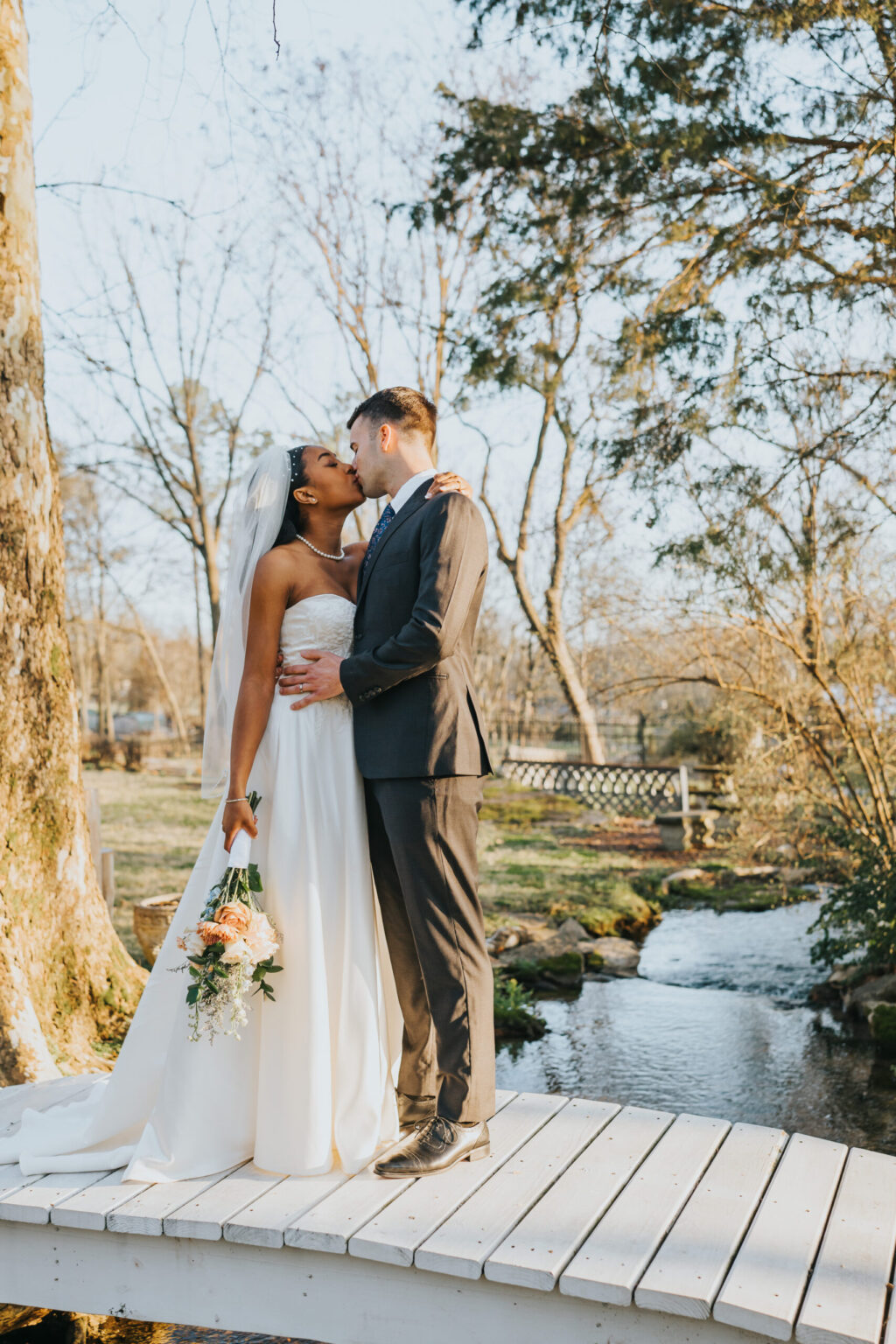 Elopement Wedding A bride and groom share a kiss on a white bridge over a small, flowing creek in a serene outdoor setting. The bride holds a bouquet of flowers and wears a white strapless gown with a veil, while the groom is dressed in a dark suit. Surrounding them are trees and greenery with a rustic fence in the background. Elopements Inc