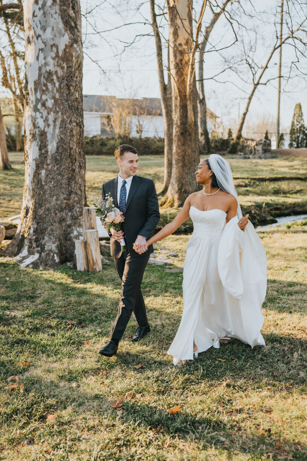 Elopement Wedding A bride and groom walk hand in hand outdoors on a sunny day. The bride is wearing a white strapless gown and a veil, holding up her dress with one hand. The groom is in a black suit with a white shirt and floral tie, holding a bouquet. They are smiling at each other with trees and grass around them. Elopements Inc