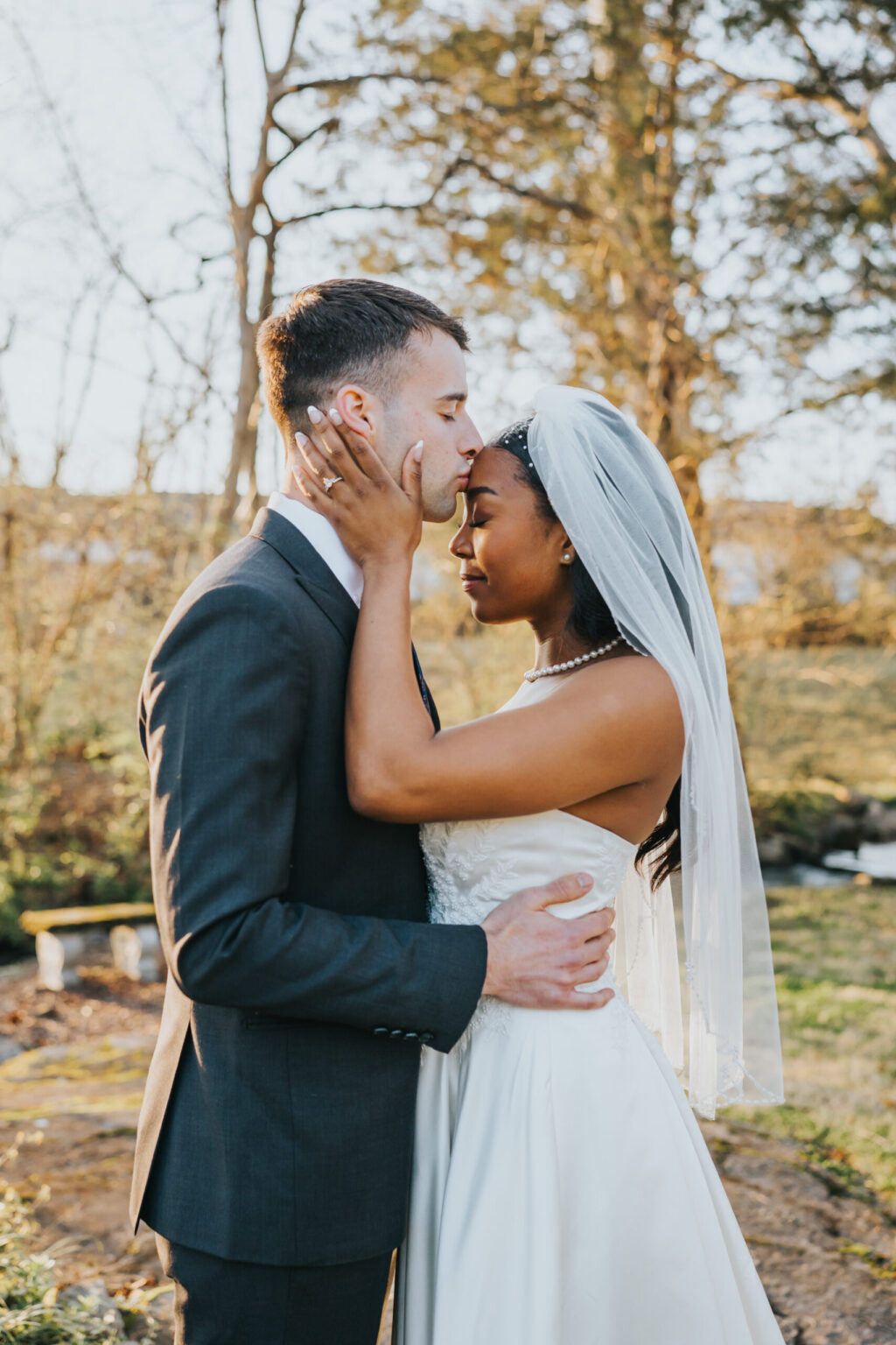 Elopement Wedding A bride and groom share an intimate moment outdoors. The groom, wearing a dark suit, gently kisses the bride, who is dressed in a white wedding gown and veil. They stand close, cradling each other's faces, with trees and soft sunlight in the background creating a serene atmosphere. Elopements Inc