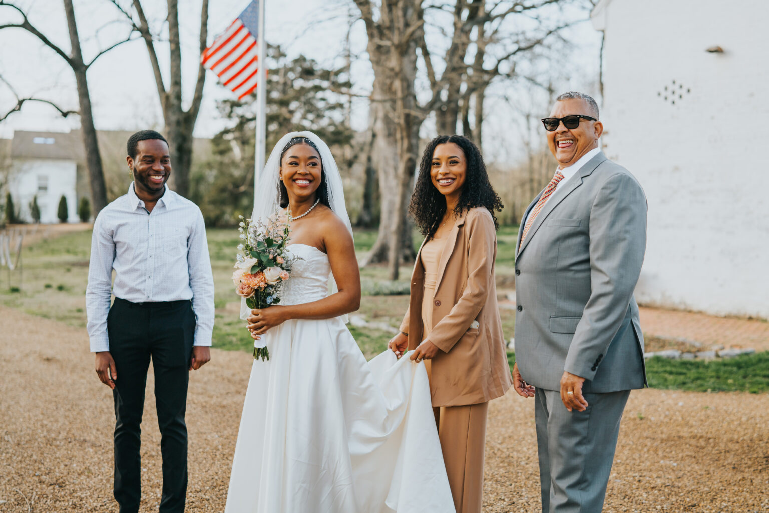 Elopement Wedding A bride in a white gown and veil holding a bouquet stands outside with three people. To her right, a woman in a tan suit holds the bride’s dress train. To her left, a man in sunglasses and a gray suit smiles, and a man in a white shirt and black pants stands smiling. An American flag is visible in the background. Elopements Inc