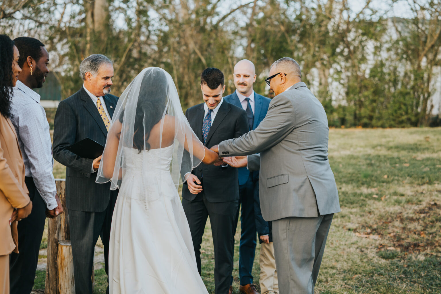 Elopement Wedding A bride and groom stand outdoors during a wedding ceremony. The bride is wearing a white gown and veil, while the groom is in a navy suit. An officiant stands to their left, and an older man in a gray suit is holding the bride's hand. Three other people are present, observing the ceremony. Elopements Inc