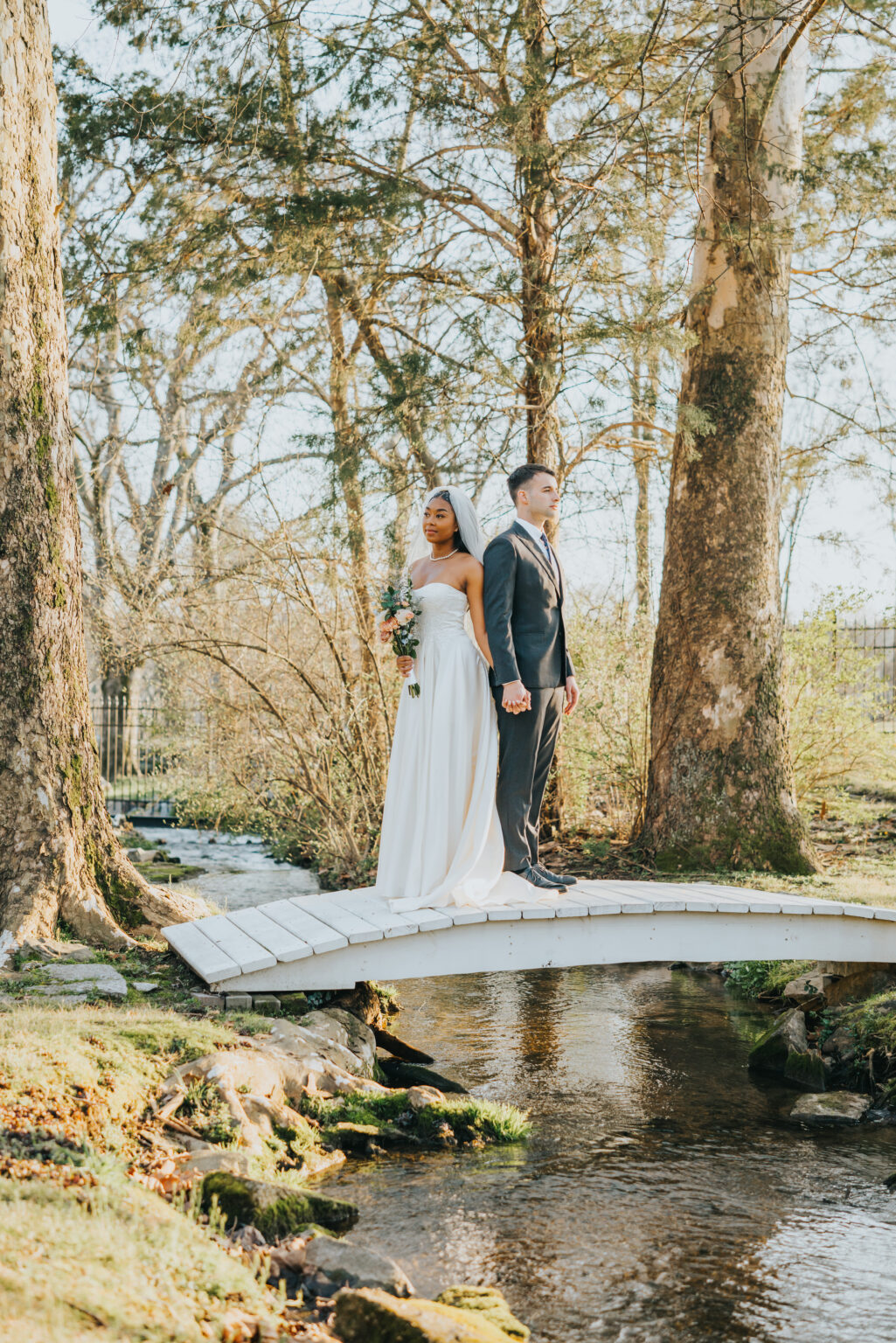 Elopement Wedding A bride and groom stand back-to-back on a small white bridge over a creek in a forested area. The bride, in a strapless white gown, holds a bouquet of flowers. The groom is dressed in a dark suit and tie. Sunlight filters through the trees, casting a serene, golden glow on the couple. Elopements Inc