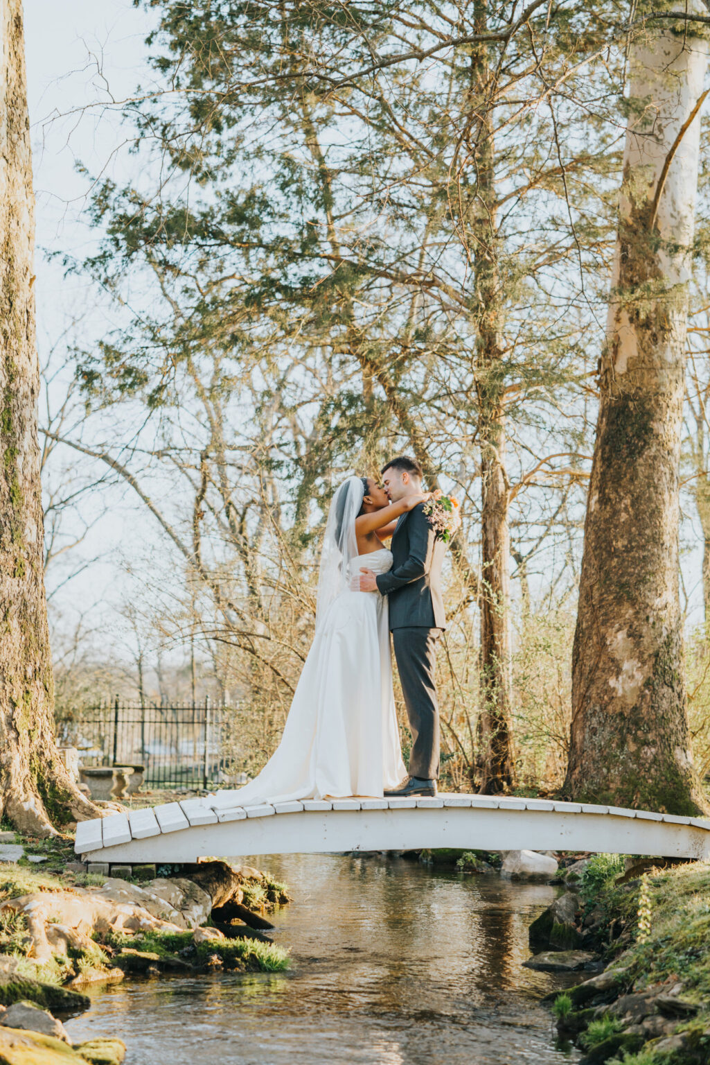 Elopement Wedding A bride and groom share a kiss while standing on a small arched white bridge over a creek. The bride is in a white dress and veil, and the groom is in a dark suit. Tall trees and a forested area surround them. The scene is serene and picturesque, with the sunlight filtering through the trees. Elopements Inc