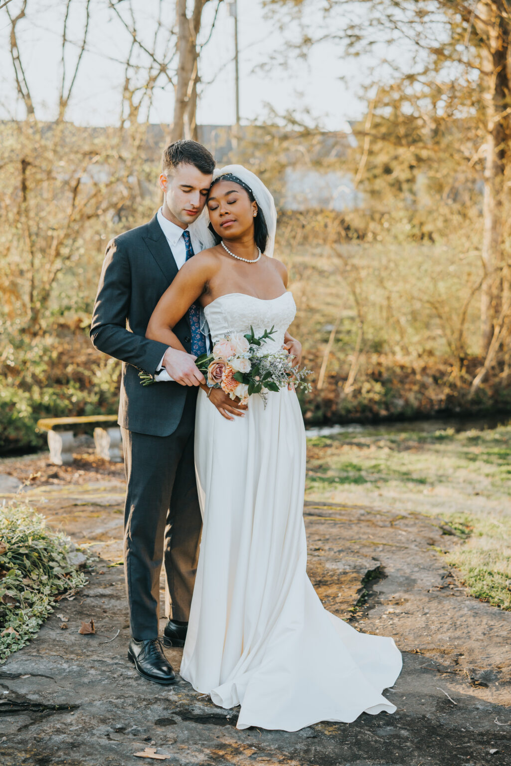 Elopement Wedding A bride and groom pose outdoors in a serene, wooded area. The groom, in a dark suit and tie, stands behind the bride, who wears a white wedding dress and veil. She holds a bouquet of pink and white flowers. Both appear content, embraced together on a stone path with greenery around them. Elopements Inc