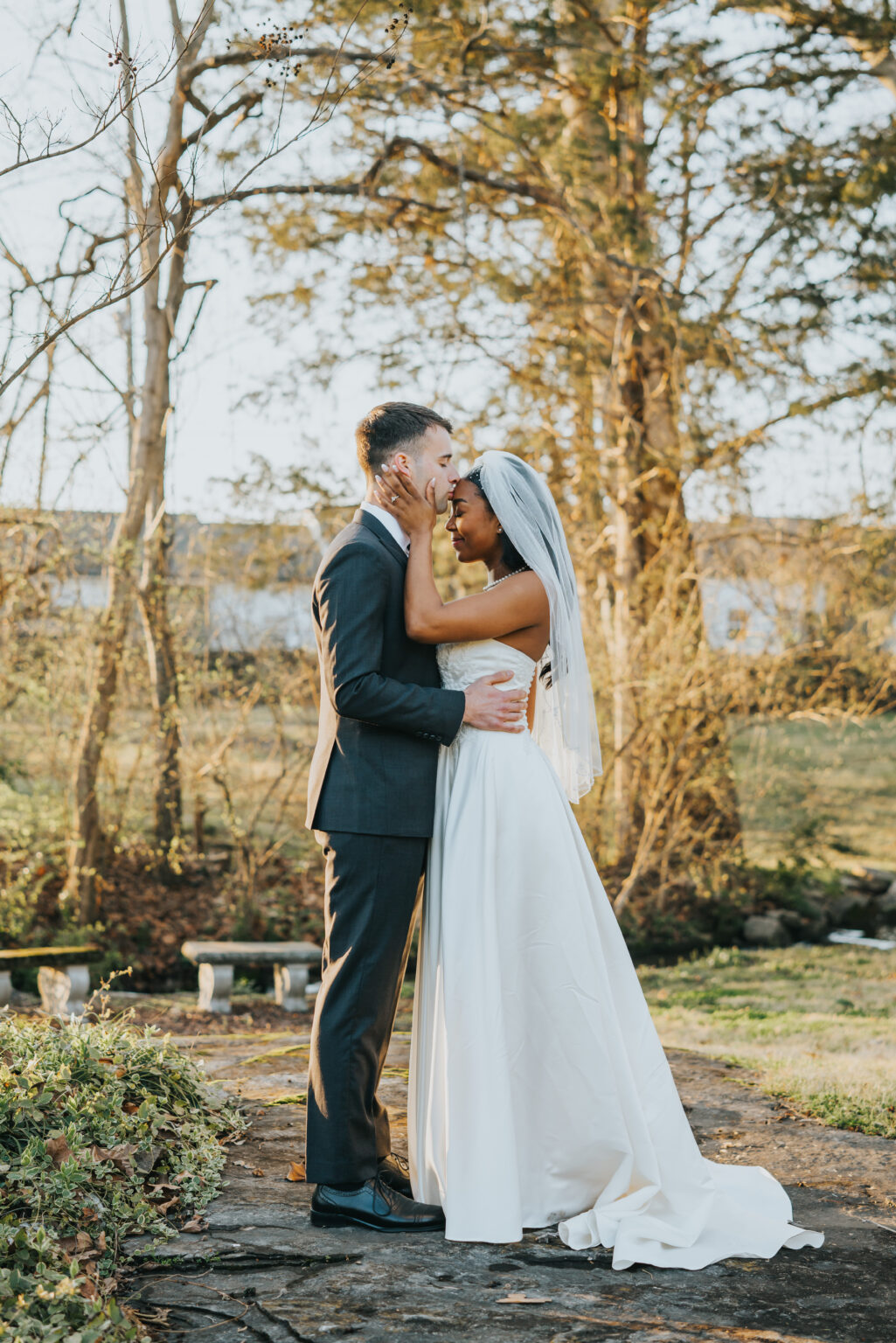 Elopement Wedding A bride and groom stand closely together in an outdoor setting with greenery and trees in the background. The bride, wearing a white dress and veil, gently cradles the groom's face. The groom, in a dark suit, has his hands on the bride's waist. They are smiling warmly at each other. Elopements Inc