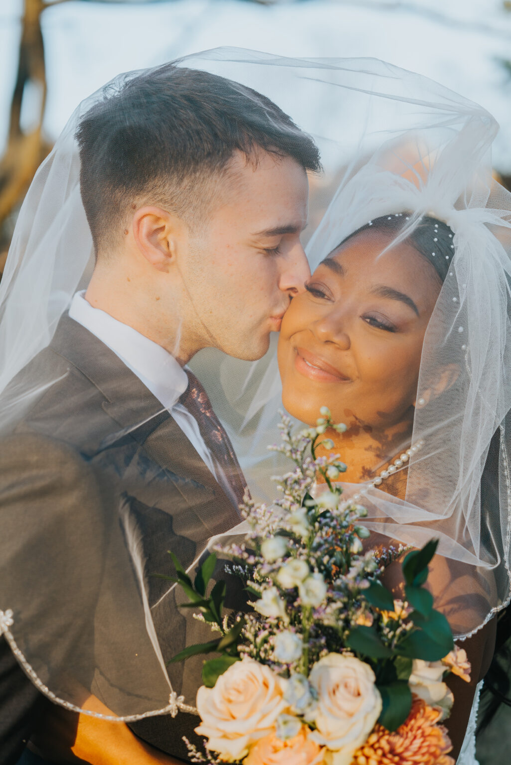 Elopement Wedding A wedding couple embraces under a veil. The groom, dressed in a dark suit, gently kisses the smiling bride's cheek. The bride wears a white veil adorned with floral embellishments and holds a bouquet with roses and greenery. They stand outdoors with sunlight casting a warm glow. Elopements Inc