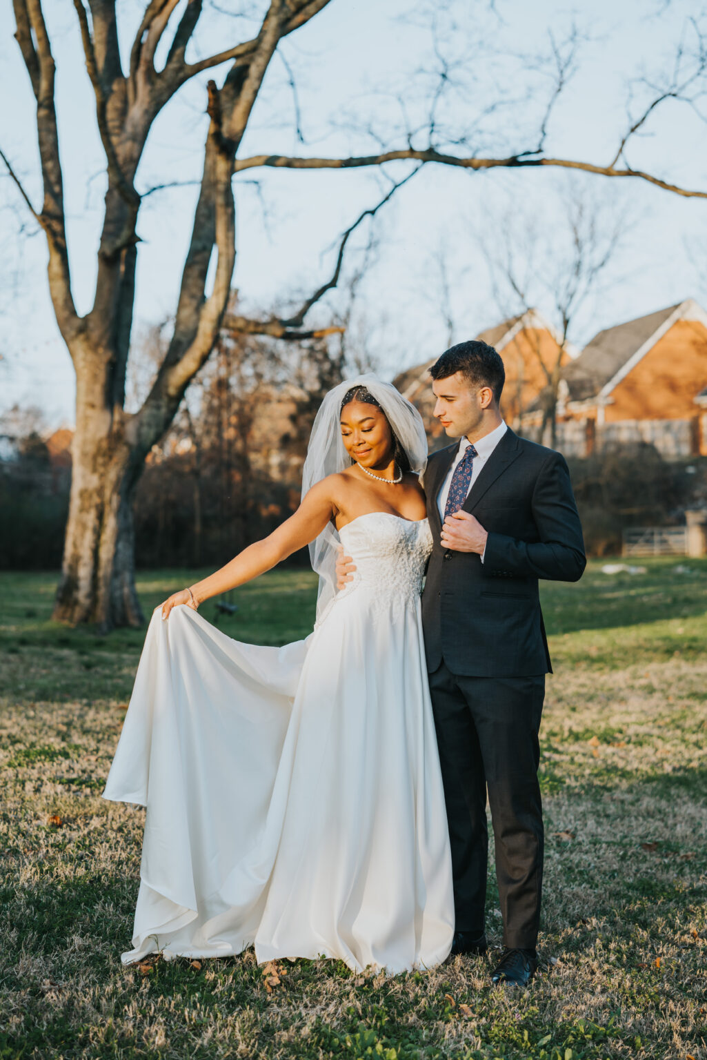 Elopement Wedding A couple in wedding attire stands in a grassy field with leafless trees in the background. The bride wears a flowing white gown and veil, holding part of her dress out. The groom is in a dark suit and tie, holding the bride's waist. They gaze at each other affectionately under a clear blue sky. Elopements Inc