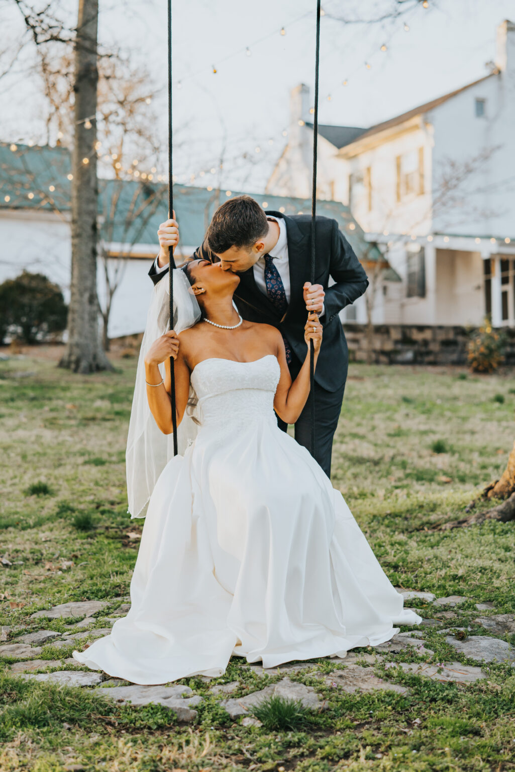 Elopement Wedding Bride in a white strapless gown and veil sits on a rustic swing, holding the ropes with both hands. Groom in a dark suit and tie leans over, kissing her passionately. They are in a grassy yard with a large house and trees adorned with string lights in the background, creating a romantic setting. Elopements Inc