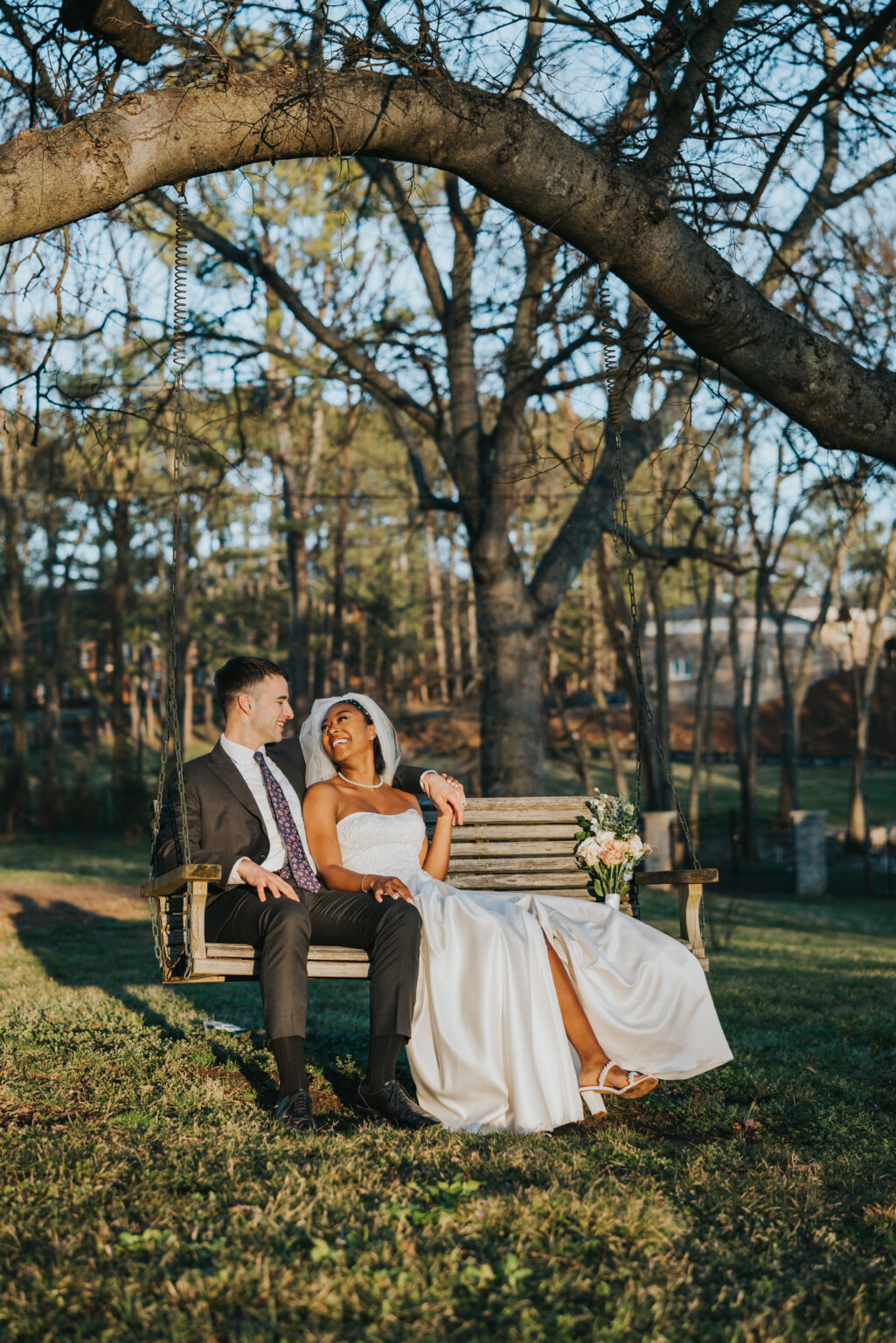 Elopement Wedding A newlywed couple sits on a rustic wooden swing hanging from a large tree in a park. The bride, in a flowing white gown and veil, joyfully leans towards the groom. The groom wears a dark suit with a patterned tie, and they both smile warmly. Sunlight filters through the trees, creating a serene background. Elopements Inc