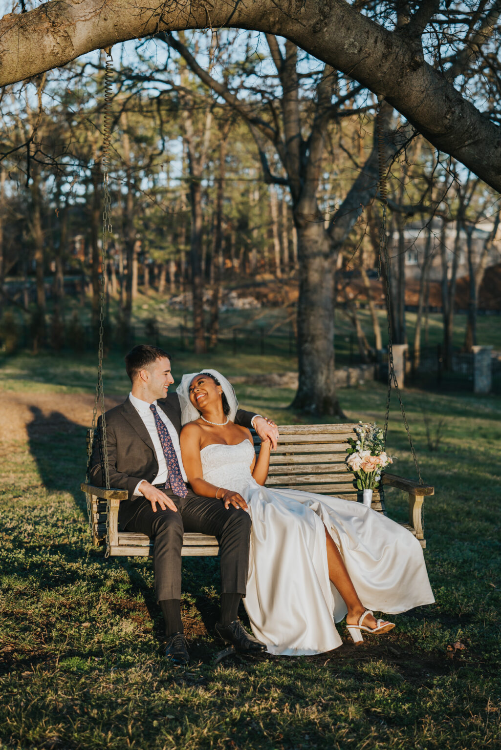 Elopement Wedding A couple sits on a wooden swing beneath a large tree in a park. The woman, dressed in a white wedding gown and holding a bouquet of flowers, looks at the man smiling. The man, wearing a gray suit and patterned tie, holds her hand. Sunlight filters through the trees, creating a serene atmosphere. Elopements Inc