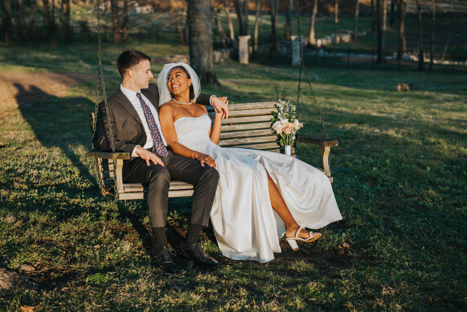 Elopement Wedding A bride and groom sit on a wooden swing outdoors, smiling at each other. The bride, in a strapless white gown and veil, holds a bouquet of flowers and extends her legs showing white sandals. The groom wears a dark suit with a patterned tie. They are surrounded by grass and trees in the background. Elopements Inc