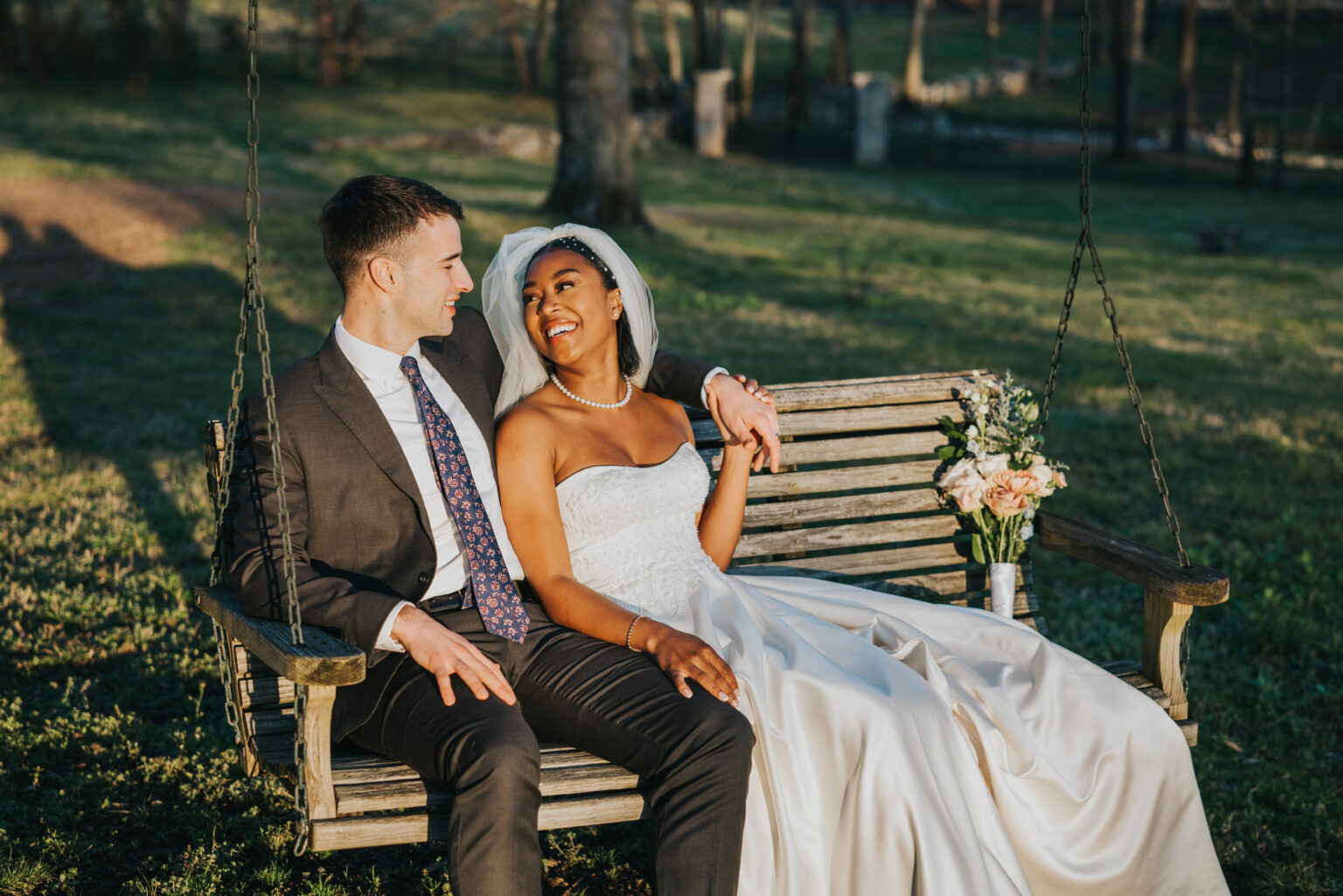 Elopement Wedding A smiling bride in a strapless white gown and veil and a groom in a dark suit with a patterned tie sit on a wooden swing. They are outdoors in a grassy area with trees visible in the background. The bride holds a bouquet of flowers, and they look joyfully at each other. Elopements Inc