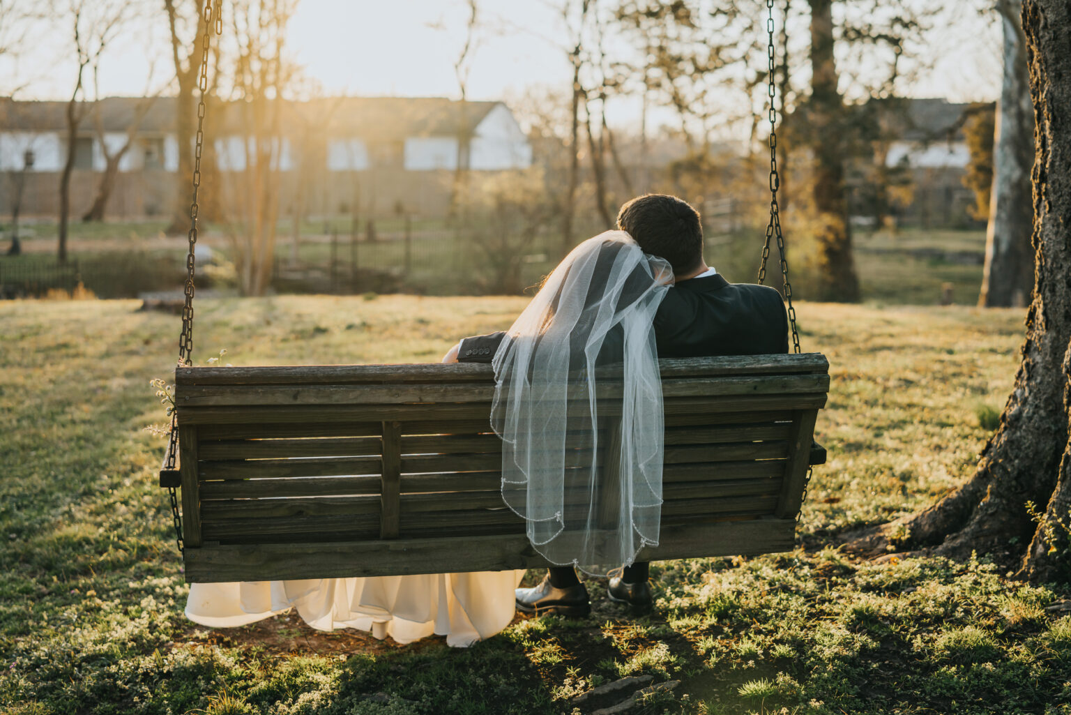 Elopement Wedding A bride and groom sit closely together on a wooden swing in a grassy yard during sunset. The bride wears a long veil and a white dress, while the groom is in a dark suit. They face away from the camera, gazing towards the horizon. Tall trees and a white building are visible in the background. Elopements Inc