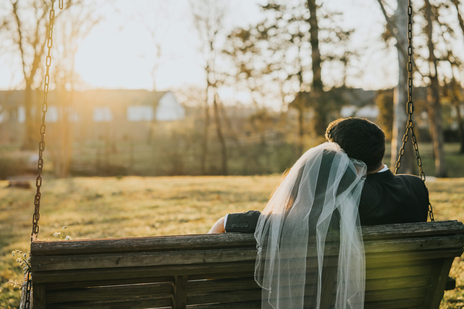 Elopement Wedding A couple sits on a wooden porch swing, viewed from behind. The person on the left wears a veil, suggesting a wedding. Sunlight streams through tree branches, casting a warm glow over the grassy yard. In the background, a fence and houses are visible under a clear sky. Elopements Inc