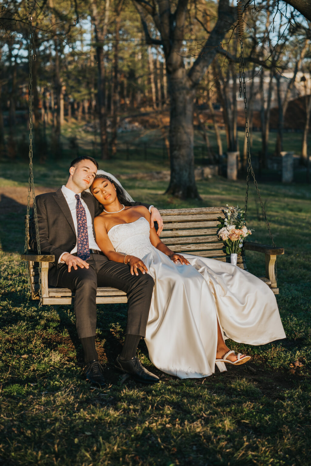 Elopement Wedding A bride in a white wedding dress and a groom in a dark suit sit closely together on a wooden swing. The bride leans her head on the groom's shoulder, and they both appear relaxed and content. The swing hangs from chains on a tree, and the background shows a grassy area with trees and sunlight filtering through. Elopements Inc