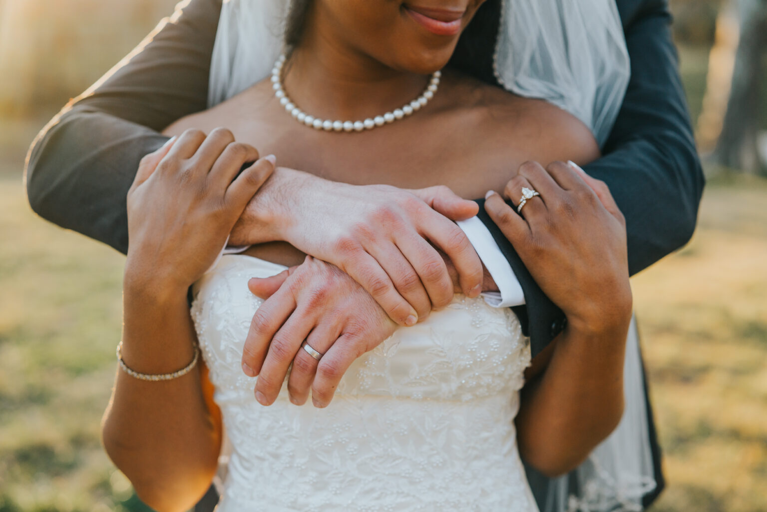 Elopement Wedding A bride and groom are embracing. The bride, in a white strapless wedding dress and pearl necklace, has her hands resting on the groom's arms, which are wrapped around her from behind. Both wear wedding rings. The background is an outdoor setting with soft natural light. Elopements Inc