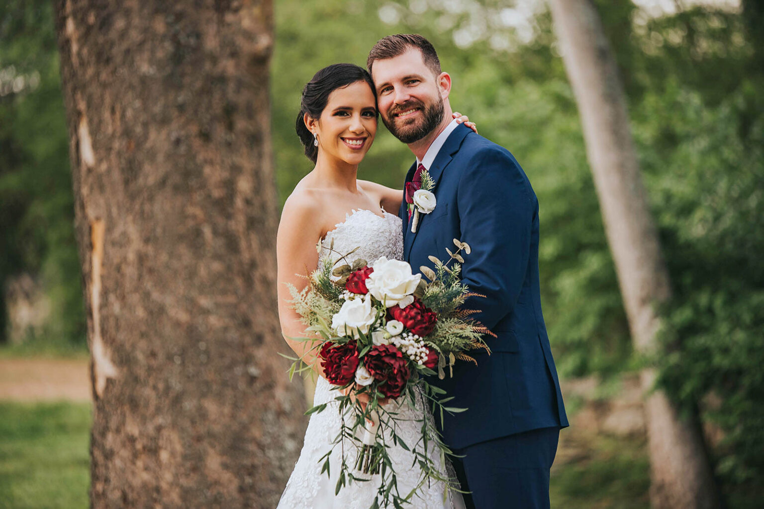 Elopement Wedding A smiling bride in a white strapless lace wedding gown holds a large bouquet of red and white flowers. The groom, wearing a navy blue suit with a maroon tie and boutonnière, stands beside her with an arm around her waist. They pose outdoors with trees and greenery in the background. Elopements Inc