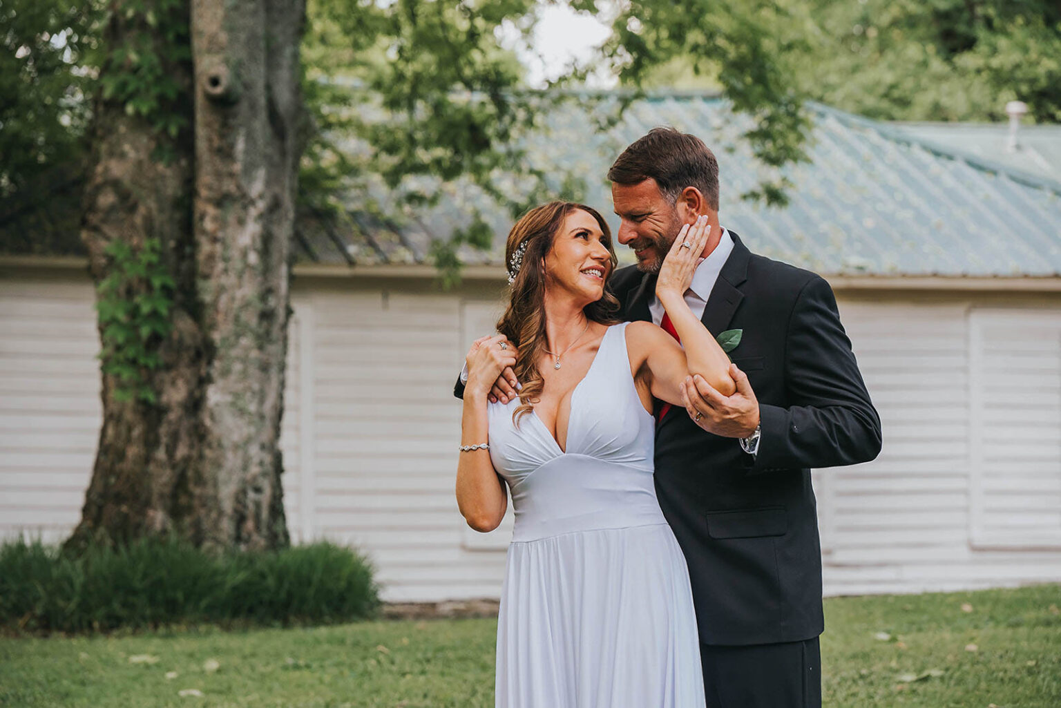 Elopement Wedding A couple stands closely in a grassy area in front of a white building with a green roof. The woman, wearing a white dress, gazes into the man's eyes while he, in a dark suit, gently holds her around the shoulders and waist. Both are smiling, creating an intimate and joyful atmosphere. Elopements Inc