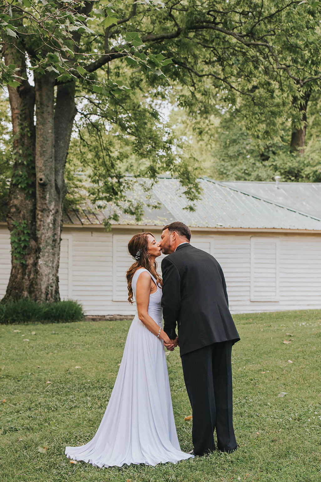 Elopement Wedding A bride and groom stand outdoors, holding hands and leaning in for a kiss. The bride wears a flowing white gown, while the groom is in a black suit. They are in front of a rustic wooden building with a tin roof, surrounded by lush green trees and grass, creating a serene, natural setting. Elopements Inc