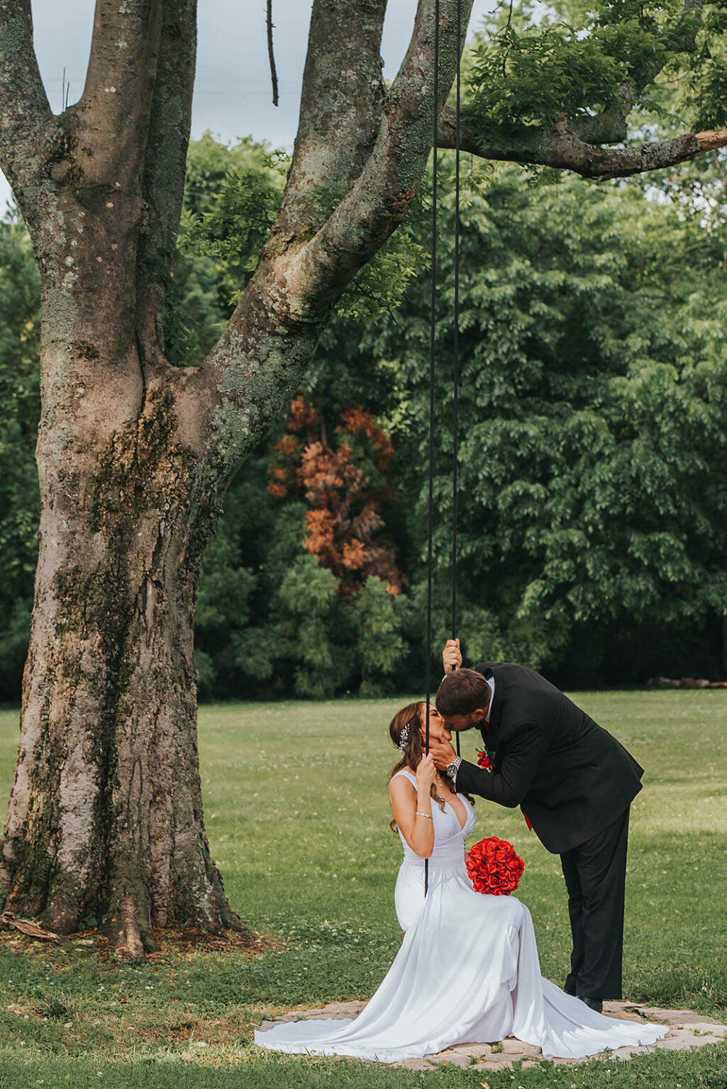 Elopement Wedding A bride in a white dress holding a bouquet of red roses sits on a swing attached to a large tree. A groom in a black suit leans down to kiss her. The background is a lush green park with various trees and grass. The scene is romantic and serene. Elopements Inc