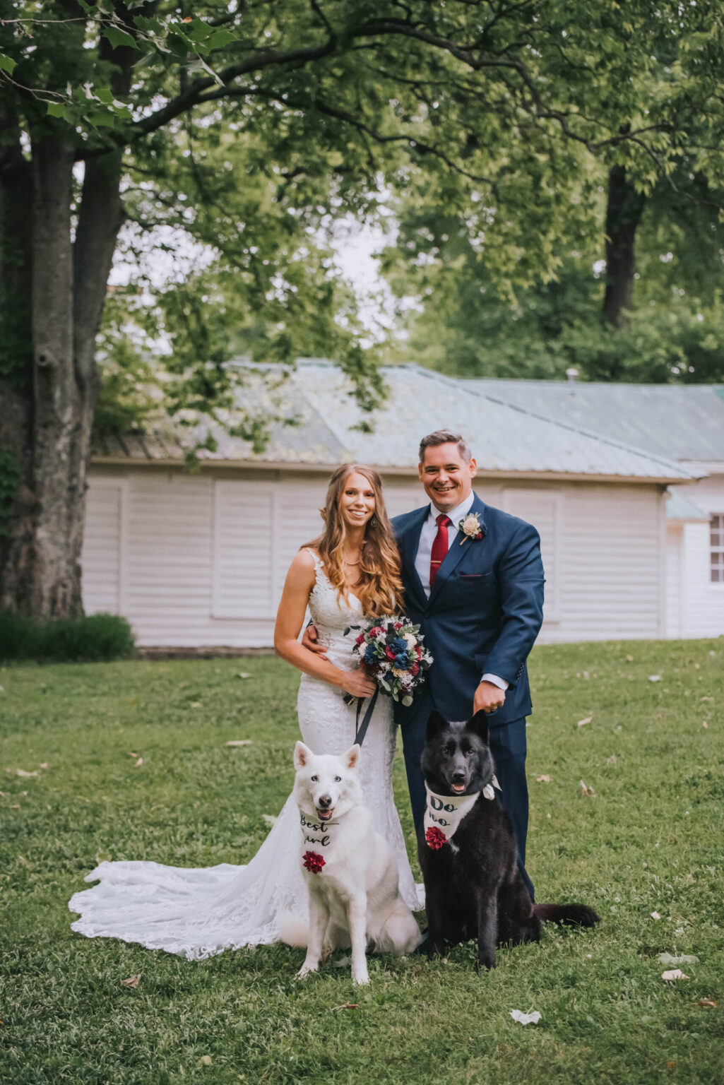 Elopement Wedding A bride in a white lace dress and a groom in a blue suit stand smiling next to each other outdoors. They hold a bouquet of colorful flowers. Two dogs with floral collars and signs reading "Dog of Honor" and "Best Dog" sit in front of them on the grass. Trees and a white building are in the background. Elopements Inc