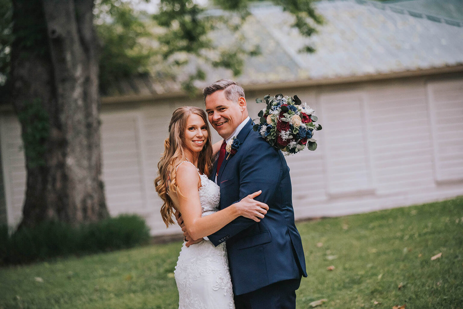 Elopement Wedding A bride in a white lace wedding dress and a groom in a navy blue suit stand outdoors, embracing and smiling in front of a white wooden building with a tin roof. The bride holds a bouquet of dark and light flowers, while the groom's arm wraps around her waist. Trees and greenery surround them. Elopements Inc