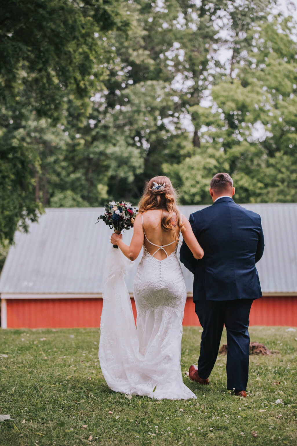 Elopement Wedding A bride in a white, backless lace wedding dress and a groom in a dark navy suit walk hand in hand on a grassy area. The bride holds a bouquet of flowers in her right hand. Behind them is a red building with a gray metal roof, surrounded by tall, leafy green trees. Elopements Inc